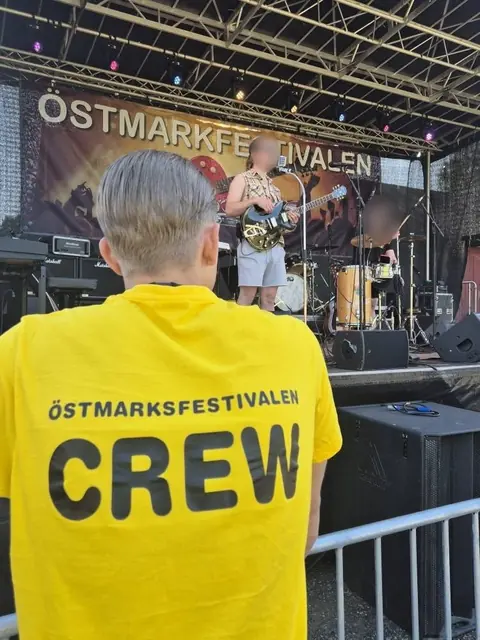 Funktionär i gul crewtröja framför scenen där ett band spelar.